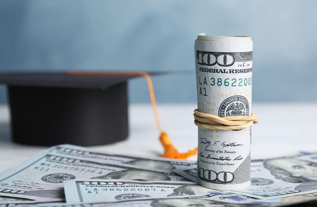 Dollar Banknotes And Student Graduation Hat On White Wooden Table Against Blue Background. Tuition F